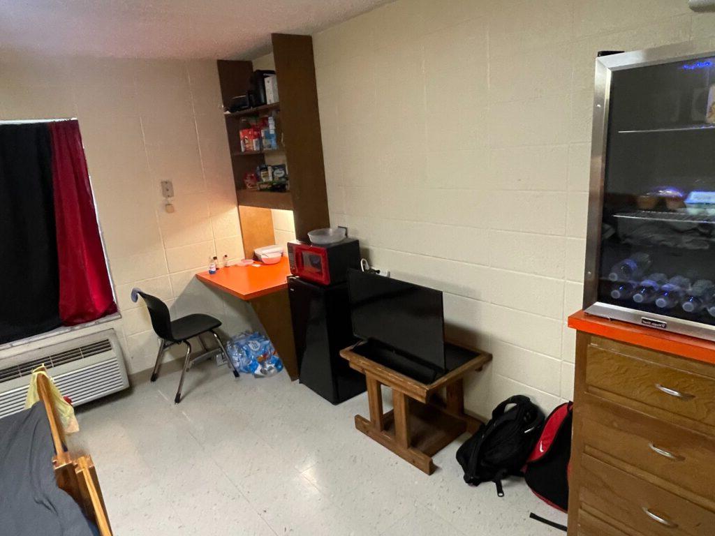 A small dorm room featuring a mini-fridge, microwave, desk with chair, TV on a stand, and various personal items. A backpack is on the floor, and a window with a curtain is partially visible.