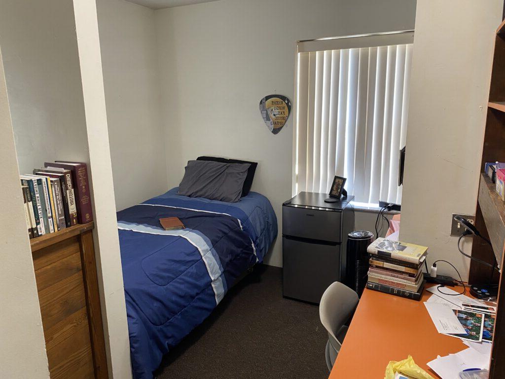 A small bedroom with a single bed, a mini fridge, a desk with books and papers, and a shelf of books. Vertical blinds cover the window.