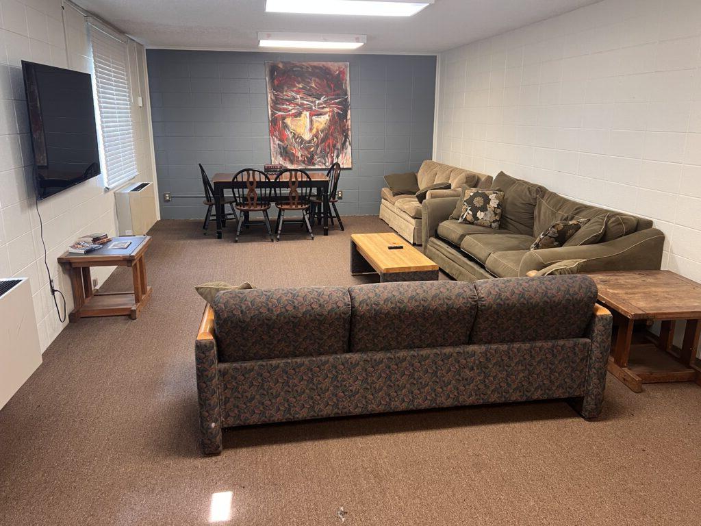 A living room with two sofas, a coffee table, side tables, a wall-mounted TV, and a dining table with six chairs. A colorful abstract painting hangs on the wall behind the dining table.