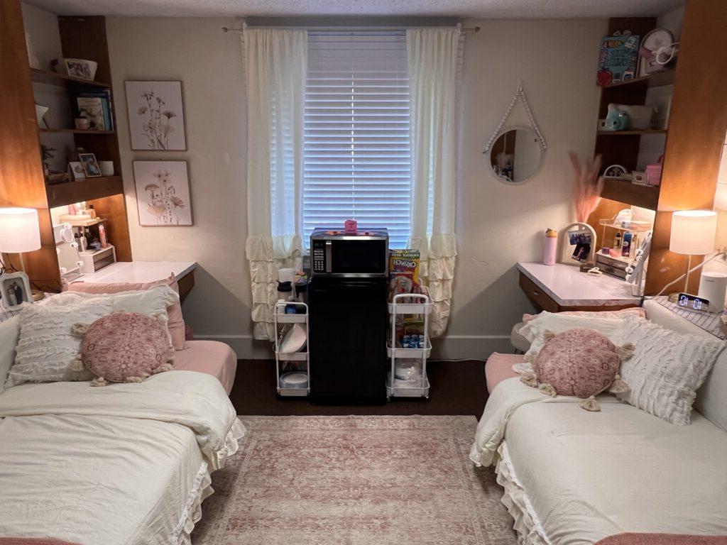 A symmetrical dorm room with two twin beds, each accompanied by a desk and shelves. A window with a blind is centered between the beds, and a small refrigerator and microwave stand below the window.