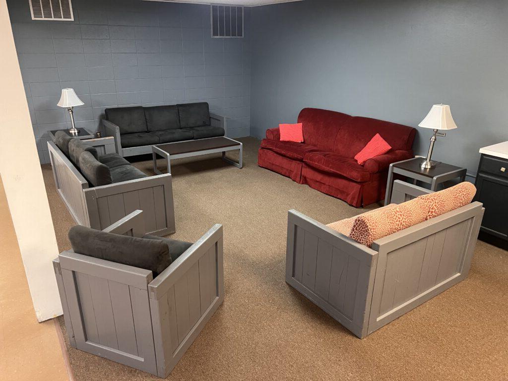 A room with a brown carpet, gray walls, and six upholstered seats arranged in a square formation around a central space, two lamps on tables beside the red sofa, and minimal additional furnishings.