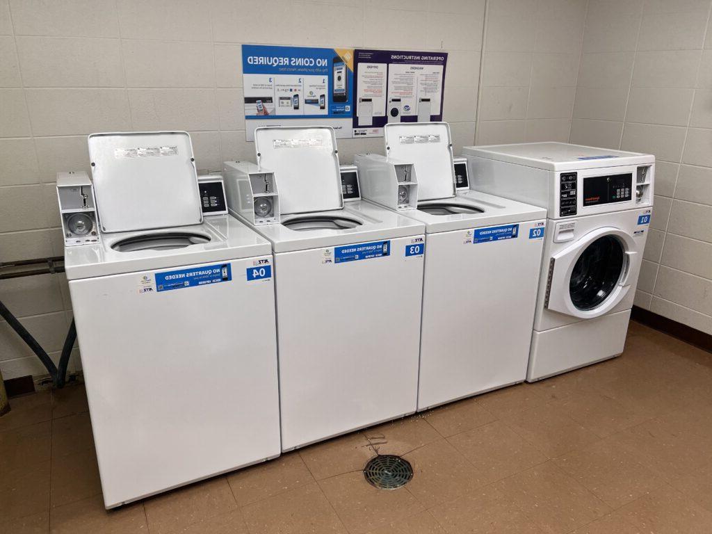 A laundromat with three top-loading washing machines and one front-loading washing machine, with instructions on a wall sign above, indicating that no coins are required.