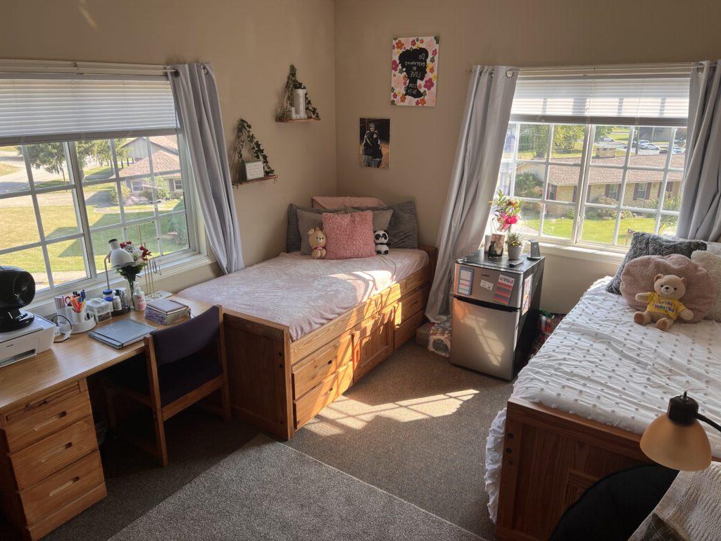 A tidy bedroom with two single beds, two desks, decorations on the walls, and natural light from two windows illuminating the room.