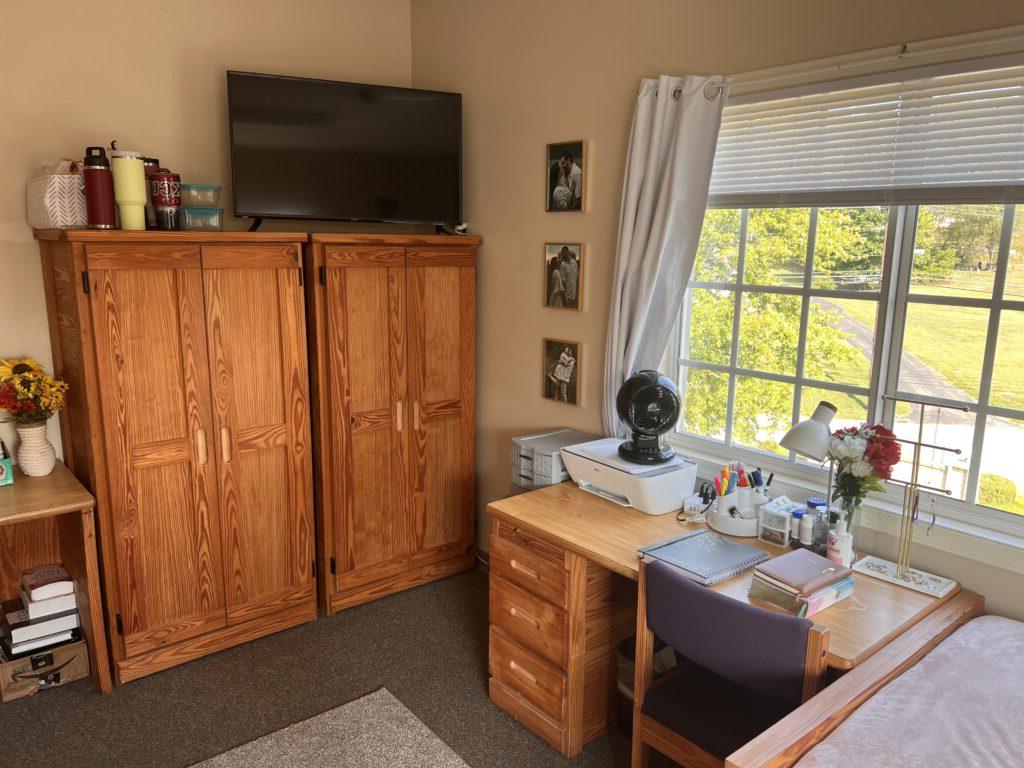 A well-organized room with a wooden desk and chair, a wooden wardrobe, a TV mounted on top of the wardrobe, and a window with white blinds. Various items are neatly arranged.